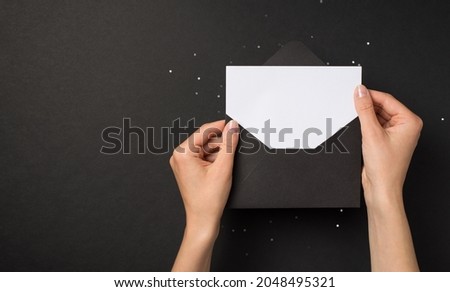First person top view photo of hands holding black open envelope with white card over sequins on isolated black background with blank space