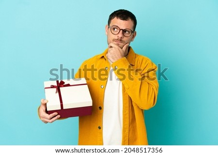 Brazilian man holding a gift over isolated blue background having doubts and thinking
