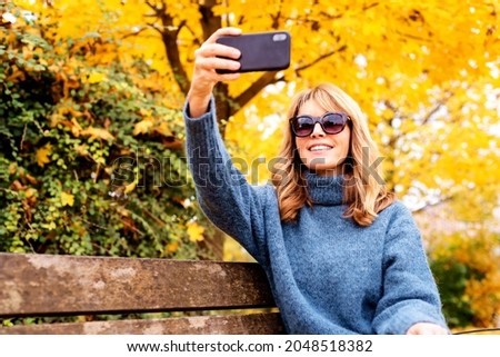 Shot of happy woman sitting on the bench in the park and taking a self portrait. Fall weather. 