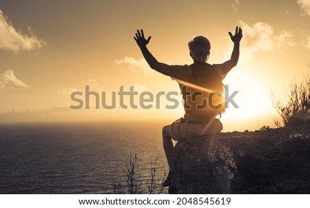 Older male hiker sitting on the edge of a mountain lifting up his arms up feeling free and inspired.  Royalty-Free Stock Photo #2048545619
