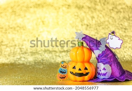 Concept photography depicting an halloween jack o lantern pumpkin head and a purple witches hat with funny ghost smiling and cardboard cutout bats against a shiny golden background.