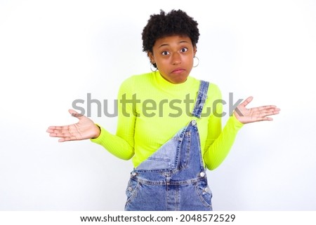 Puzzled and clueless young American woman with short hair wearing denim overall against white wall with arms out, shrugging shoulders, saying: who cares, so what, I don't know. Negative human