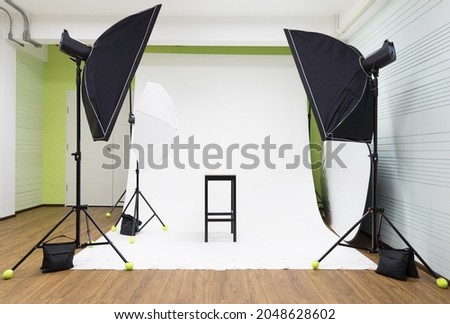 Empty nobody indoor room studio white photoshoot scene with professional photographer equipment set softbox strobe flash umbrella reflector light bulb on tripod stand with black chair ready for model.