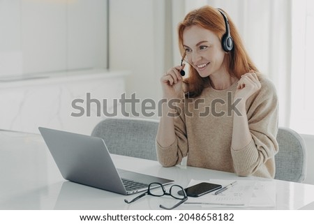 Pleasant smiling redhead woman in headset communicating with colleagues during video call on laptop computer, female freelancer participating in online meeting while working remotely from home Royalty-Free Stock Photo #2048658182