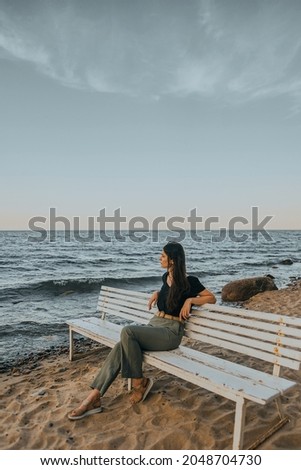 Calm girl with dark hair is sitting on the white bench on the seashore.