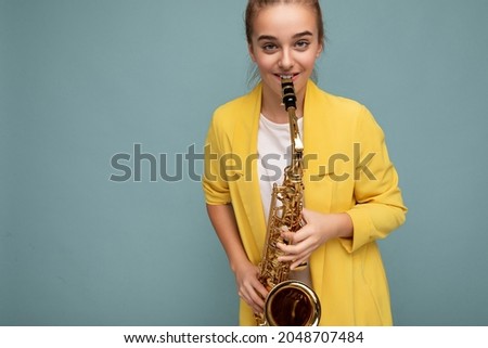 Photo shot of beautiful happy smiling brunette little girl wearing stylish yellow jacket standing isolated over blue background wall playing saxophone looking at camera