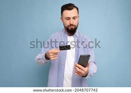 Handsome man wearing everyday clothes isolated on background wall holding and using phone and credit card making payment looking at smartphone screen
