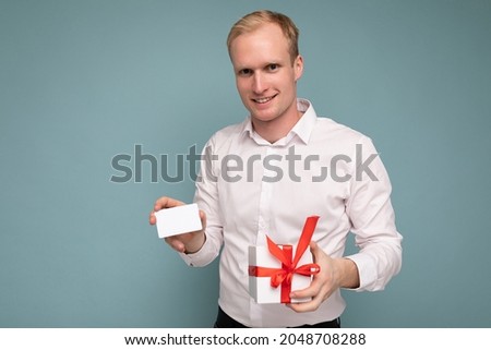 Photo shot of handsome positive smiling young blonde male person isolated over blue background wall wearing white shirt holding white gift box with red ribbon and credit card looking at camera