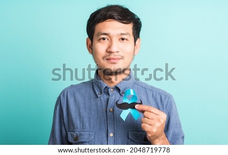 Asian portrait happy handsome man posing he holding a light blue ribbon and mustache, studio shot isolated on blue background, Prostate Cancer Awareness in November concept