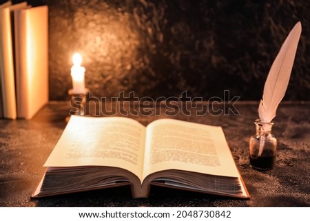 Open old book with feather and burning candle on table against dark background