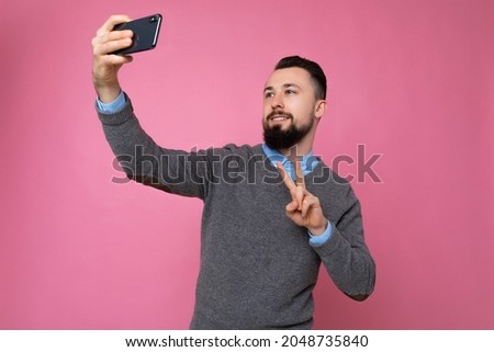 Photo of positive handsome young brunette unshaven man with beard wearing casual grey sweater and blue shirt isolated on pink background wall holding smartphone taking selfie photo looking at mobile