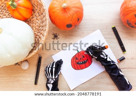 Preparation for the holiday Halloween. Hands in black gloves with bones are drawing a sketch with markers a laughing pumpkin, on a wooden desk surrounded by pumpkins and spiders, top view.