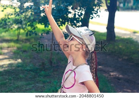 Adorable little girl with long braided hair in baseball cap outdoor on sunny day landscape. Youth culture lifestyle authentic photo. Happy traveler child on green grass in park. School kid walking.
