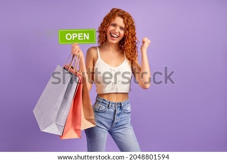 Cute red-haired girl with curls and shopping bags holding a sign with the inscription open isolated on a purple background. Shopping concept, shopaholism, sale.