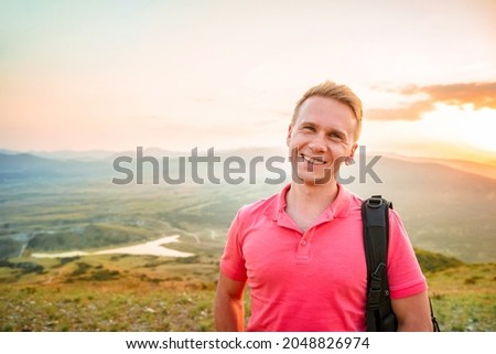 A young blond man in a pink shirt conquered the top of the mountain at sunset. Beautiful mountain landscape with a gradient. The concept of enjoying nature.