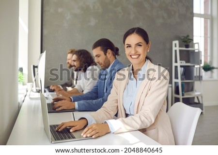 Business people using modern computers in office. Portrait of happy cheerful beautiful young woman sitting at table with team of colleagues, working on project on laptop, looking at camera and smiling