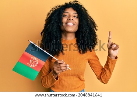 African american woman with afro hair holding afghanistan flag smiling with an idea or question pointing finger with happy face, number one 