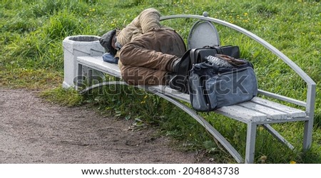 an adult homeless tramp sleeps on a park bench in the open air next to a bag and briefcase. Royalty-Free Stock Photo #2048843738
