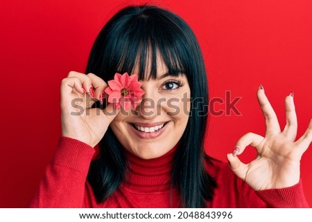 Young hispanic woman holding sunflower over eye doing ok sign with fingers, smiling friendly gesturing excellent symbol 