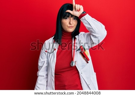 Young hispanic woman wearing doctor uniform and stethoscope making fun of people with fingers on forehead doing loser gesture mocking and insulting. 
