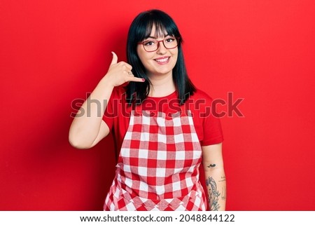 Young hispanic woman wearing cook apron and glasses smiling doing phone gesture with hand and fingers like talking on the telephone. communicating concepts. 