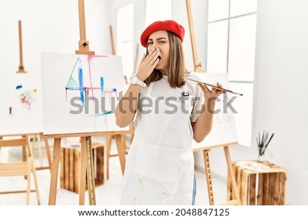 Young artist woman painting on a canvas at art studio bored yawning tired covering mouth with hand. restless and sleepiness. 