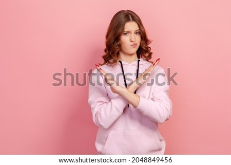 Portrait of serious teen girl with brunette hair in hoodie making x sign with crossed hands, gesturing stop, warning to ban, body language. Indoor studio shot isolated on pink background