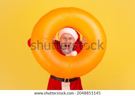 Portrait of elderly man with gray beard wearing santa claus costume has amazed shocked expression, celebrating winter holidays in warm resort. Indoor studio shot isolated on yellow background.