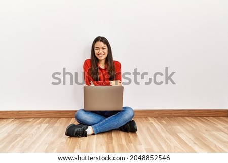 Young brunette woman sitting on the floor at empty room with laptop happy face smiling with crossed arms looking at the camera. positive person. 
