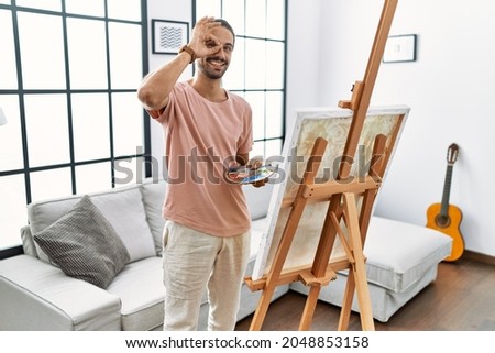 Young hispanic man with beard painting on canvas at home doing ok gesture with hand smiling, eye looking through fingers with happy face. 