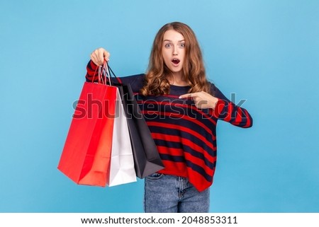 Surprised woman wearing striped casual style sweater, pointing at shopping bags, looking at camera with open mouth and shocked facial expression. Indoor studio shot isolated on blue background.