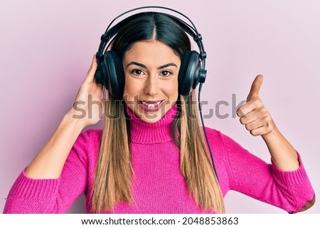 Young hispanic woman listening to music using headphones smiling happy and positive, thumb up doing excellent and approval sign 