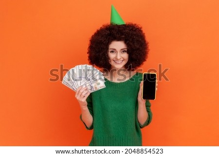 Happy woman with Afro hairstyle wearing green casual style sweater holding fan of dollar bills and showing phone with empty display. Indoor studio shot isolated on orange background.
