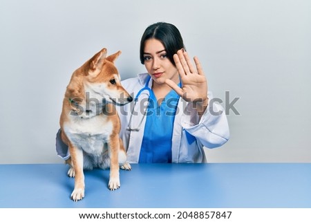 Beautiful hispanic veterinarian woman checking dog health with open hand doing stop sign with serious and confident expression, defense gesture 