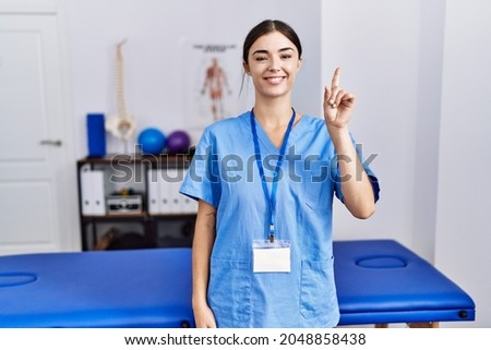 Young hispanic woman wearing physiotherapist uniform standing at clinic showing and pointing up with finger number one while smiling confident and happy. 