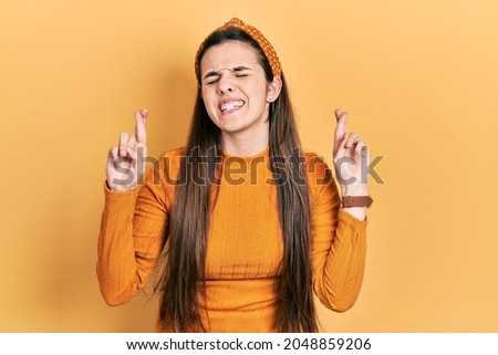 Young brunette teenager wearing casual yellow sweater gesturing finger crossed smiling with hope and eyes closed. luck and superstitious concept. 