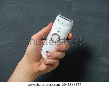 Electric epilator in white color in a man's hand on a gray textured background. Means for hair removal, beauty care, beauty