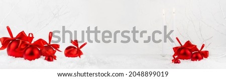 Christmas festive banner in white and red color - white burning candles, heap of rich red shimmer balls with satin ribbon, bows in snowd under white frosty branches in winter fores, copy space, frame.