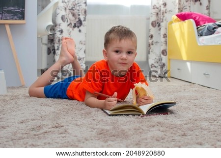 A little boy draws on the floor and eats a banana in his room. A happy child draws with pencils, lying on the floor of the house. Children's leisure. Family and children.