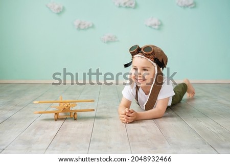 A little boy in aviation helmet lies on the floor next to a wooden toy plane and smiles. The boy dreams of becoming a pilot. Royalty-Free Stock Photo #2048932466