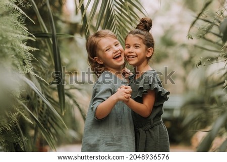 Two cute smiling little girls belonging to different races, in linen clothes, holding hands and walking in the botanical garden. Children explore tropical plants and flowers in the greenhouse. Royalty-Free Stock Photo #2048936576
