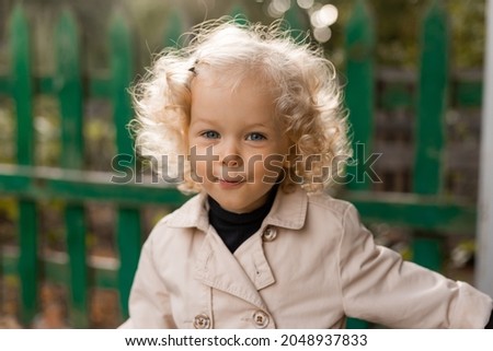 portrait of a cute little curly blonde girl with blue eyes in a white linen raincoat stands against the background of an old fence at sunset. lifestyle. space for text. High quality photo