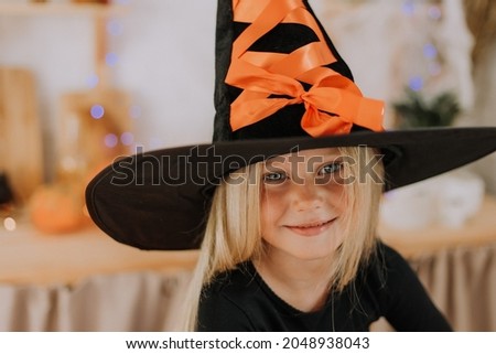 close-up portrait of a little blonde girl with blue eyes in a black hat and a black sweater in the Halloween decorations. witch costume. High quality photo
