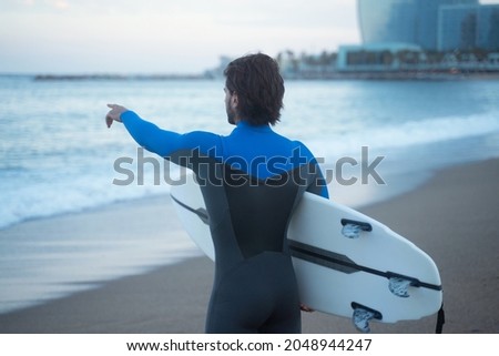 From the back silhouette shot of surfer standing at beach and holding surf board, pointing at waves with hand