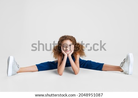 Portrait of little cute curly girl in blue stylish jumpsuit sitting on a twine and having fun isolated on white studio background. Childhood, education, emotions, sport, healthy lifestyle concept, ad