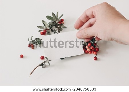 Woman hand holding paper business card. Red lingonberries fruit with green leaves on white table background. Winter wedding stationery mockup scene. Selective focus, blurred background. Healthy herb.