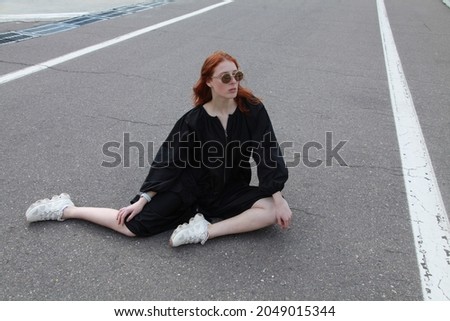 Full length urban portrait of ginger female model wearing oversize fashionable black raincoat and sneakers while sitting on the road