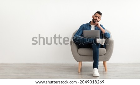 Thoughtful young Arab guy using laptop, sitting in armchair, looking aside at empty space against white studio wall, banner. Young Eastern man working or studying online, thinking over something Royalty-Free Stock Photo #2049018269