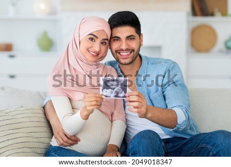 Happy pregnant muslim couple demonstrating baby sonography photo and smiling to camera, resting together at home. Loving arab cpouses enjoying future parenthood