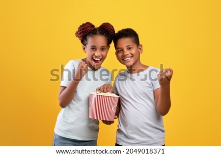 Cheerful brother and sister watching cartoon or nice movie together at cinema, eating popcorn, raising clenched fists up, yellow studio background, kids entertainment concept